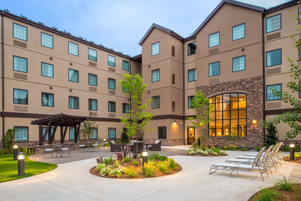 A beautifully landscaped courtyard with lush gardens surrounds a welcoming seating area. Large windows from the hotel overlook the outdoor space, while hints of morning sunlight or sunset glow create a warm, inviting atmosphere. The courtyard feels ideal for families relaxing together or unwinding after a day of exploring local attractions.