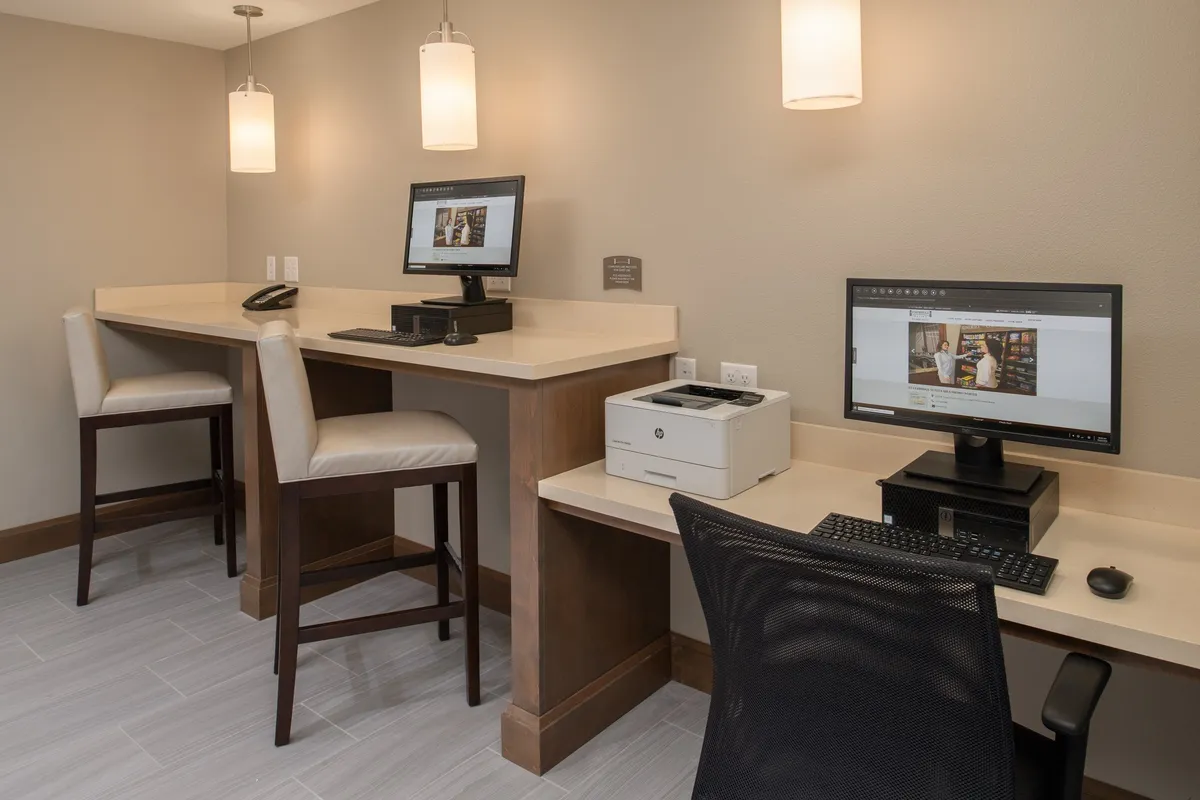 A professional business center with workstations, computers, a printer, and stylish bar stools beneath pendant lighting. The space is designed for productivity, offering a quiet environment for guests to work or conduct virtual meetings at any time of day.