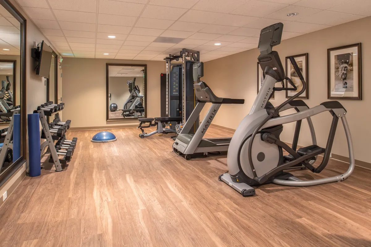 A bright, well-equipped fitness center featuring an elliptical machine, weight bench, free weights, exercise balls, and mirrored walls. The space is designed for both individual workouts and small group fitness sessions, with plenty of natural light and motivational ambiance.