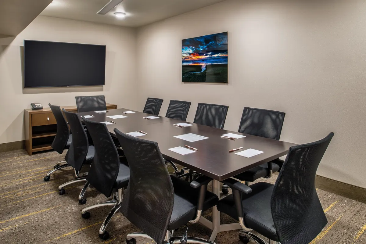 A modern workspace with a large conference table, comfortable office chairs, a TV screen, and artwork on the wall. The room is designed to foster productivity in a calm, well-lit environment. The decor is sleek and contemporary, offering a professional vibe.