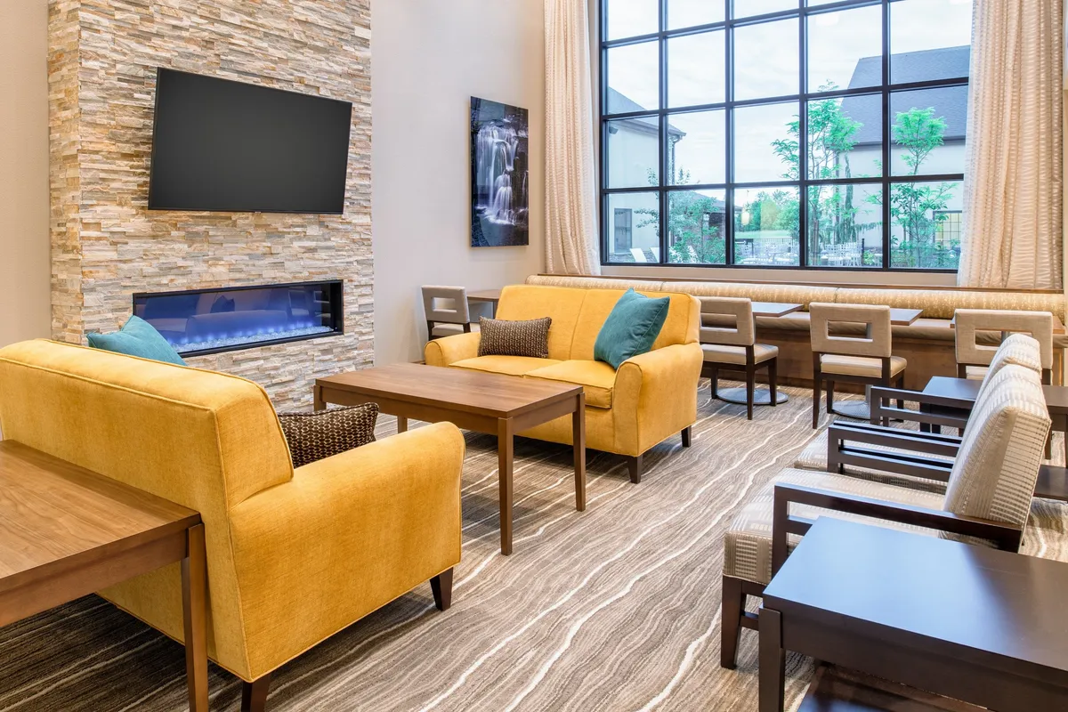 A stylish living area featuring cheerful yellow sofas, wooden tables, a large window letting in natural light, and a cozy fireplace. The setting feels warm and inviting, suitable for both relaxation and casual socializing. The space looks bright and comfortable, suggesting a restful environment.