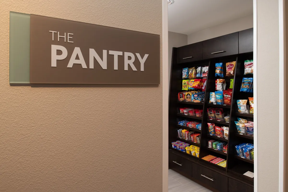 A welcoming pantry area inside the property, featuring prominent signage reading 'THE PANTRY.' Shelves are filled with snacks and food items, inviting guests to browse. The mood is relaxed, and the space encourages interactions and quick snack selections.