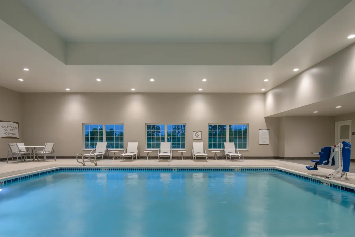 This image features a bright indoor swimming pool, surrounded by inviting lounge chairs positioned near large windows that let in natural light. The calm water, soft lighting, and relaxed seating create a welcoming space for both recreation and casual socializing.