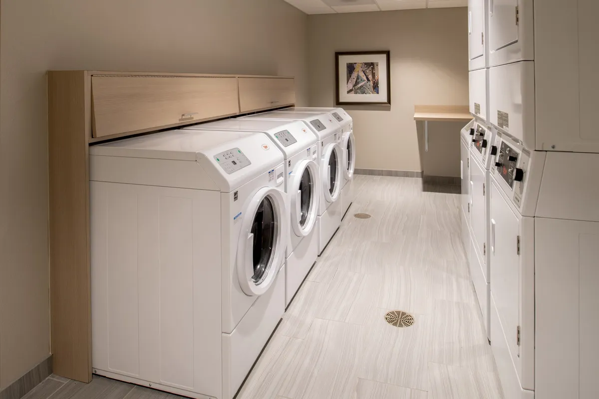 A functional and inviting laundry room with multiple washing machines and dryers, a wooden countertop for folding clothes, and framed artwork adding a touch of warmth. The setup encourages both practicality and comfort for guests on extended stays.