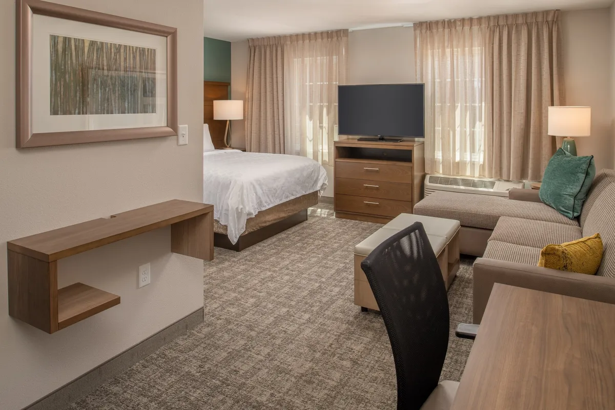 The image showcases a bright and modern studio suite with a plush Queen bed, a cozy seating area perfect for sharing a meal or relaxing, a flat-screen TV for movie nights, and a convenient desk space for catching up on work. Soft lighting and tasteful decor create an inviting, comfortable atmosphere.
